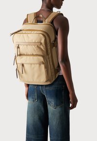 Beige Nylon-Rucksack mit mehreren Reißverschlusstaschen, verstellbaren Schulterriemen und kontrastierenden braunen Akzenten. Hat eine elegante rechteckige Form.