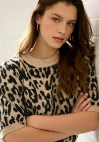 Beżowy sweter dzianinowy z czarnym nadrukiem w panterkę, z prążkowanym okrągłym dekoltem, krótkimi rękawkami i miękką teksturą. Modelka ma długie brązowe włosy i kolczyki w kształcie kół.