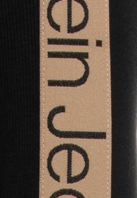 Černá tkanina s hnědým štítkem s černým textem. Štítek je našitý na okraji, což přidává kontrastní prvek do designu.