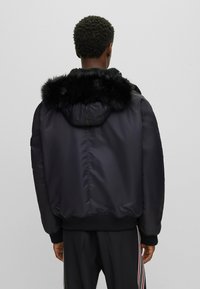 Chaqueta de plumas negra con capucha de piel sintética, puños y dobladillo acanalados. Tejido suave con un acabado ligeramente brillante, con cierre de cremallera en la parte posterior.