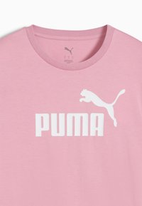 Růžové bavlněné tričko s kulatým výstřihem, na němž se nachází velké bílé logo "PUMA" a nad ním grafika skákající pumy v bílé barvě.