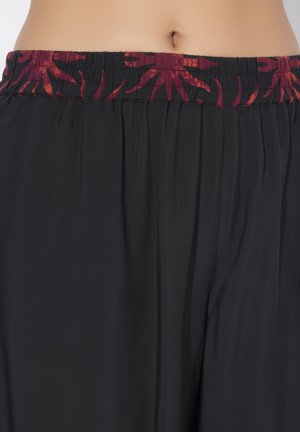 Taille en navel zichtbaar boven losse zwarte broek met brede elastische tailleband in rood-zwart abstract patroon.