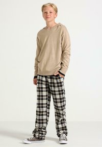 Tenåringsgutt som står med hendene i lommene, iført beige hettegenser, svart-hvite rutete bukser og grå sneakers foran en ensfarget bakgrunn.
