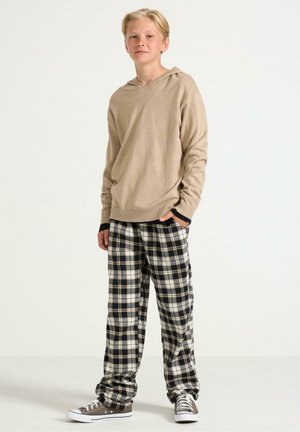 Un adolescent se tient avec les mains dans les poches, portant un sweat à capuche beige, un pantalon à carreaux noir et blanc, et des baskets grises sur un fond uni.