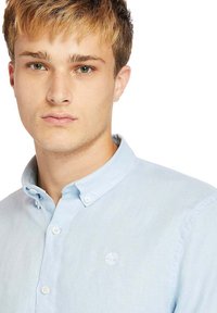 Camicetta azzurra chiara con colletto, caratterizzata da un piccolo logo bianco e chiusure a bottoni. Tessuto liscio, design casual.