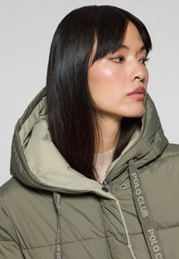 Puffig olivgrön jacka med en ljusgrå huva, med justerbara dragsnören och kviltad textur. Tyget verkar mjukt och värmande.