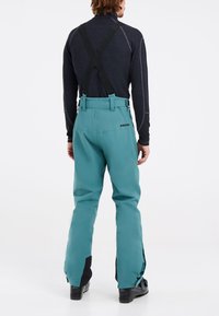 Pantalones de esquí impermeables en color turquesa con tirantes negros ajustables, cremalleras laterales y detalles negros cerca de los puños. Logo de la marca en la parte trasera.