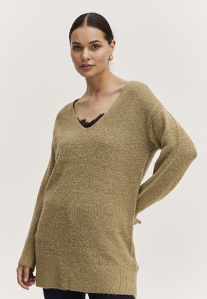 V-Ausschnitt, übergroßer Pullover in Olivgrün, strukturiertes Strickmaterial mit langen Ärmeln und Seitenschlitzen, über einem Oberteil mit schwarzem Spitzendetail getragen.