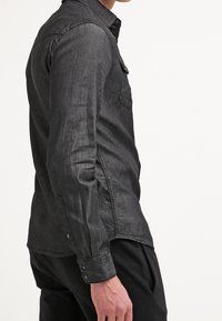 Ciemnoniebieska koszula dżinsowa z kołnierzykiem zapinanym na guziki, długimi rękawami, kieszeniami na piersi i delikatną teksturą, zaprezentowana z bocznego widoku.