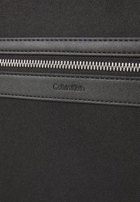 Gros plan sur un sac en tissu noir avec un matériau en toile texturée, un accent en cuir noir lisse et une fermeture éclair en métal argenté portant le logo Calvin Klein.
