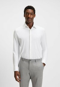 Chemise blanche à manches longues avec un col étalé, design à boutons sur le devant et texture lisse, associée à un pantalon ajusté gris clair.
