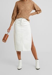 Gonna midi bianca in denim con spacchi alti, abbinata a un maglione lavorato marrone e stivaletti bianchi. Gli accessori includono una borsa marrone.