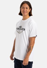 Camiseta de algodón blanca con un logo negro de Quiksilver y texto en el frente. Mangas cortas, cuello redondo y ajuste relajado.