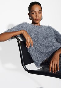 Fluffy, short-sleeve, light gray top with a textured finish; worn over dark denim, model seated on a black chair. Silver hoop earrings visible.