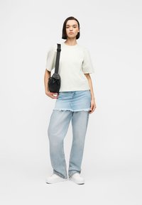 Νεαρή γυναίκα που φοράει λευκό πουκάμισο με φουσκωτά μανίκια, στρωτή φούστα από ανοιχτό τζιν πάνω από τζιν παντελόνι, λευκά αθλητικά παπούτσια και μαύρη τσάντα ώμου, στέκεται σε λευκό φόντο.