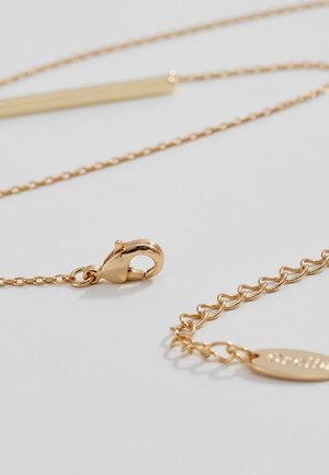 Gros plan d’un collier en chaîne dorée avec un fermoir de type homard et une chaîne réglable se terminant par une petite plaque ovale.