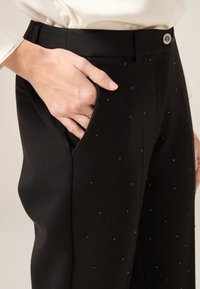 Czarne spodnie o gładkiej fakturze, z delikatnymi kwadratowymi zdobieniami i standardowym paskiem. Kieszeń widoczna z boku.