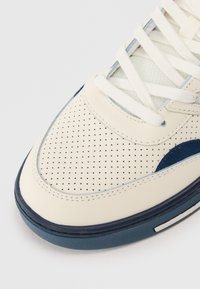 Baskets en cuir crème avec un design perforé, des accents bleu marine et des lacets blancs. Bout rond, semelle rembourrée et détails cousus.