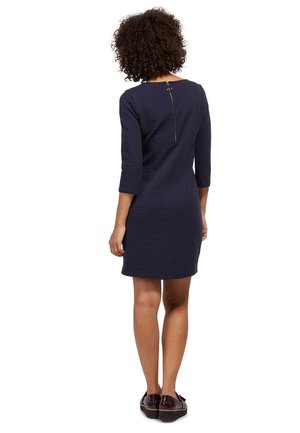 Marineblå knælængde kjole med tekstureret stof, trekvartærmer og lynlåslukning bagpå. Båret med mørke slip-on sko.