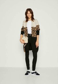 Cardigan leopardato in beige e nero, abbinato a una maglietta bianca e leggings neri. La modella tiene una piccola borsa color crema e indossa sneakers nere.
