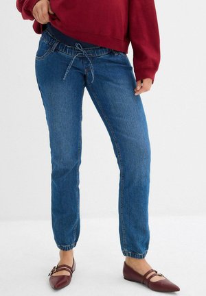 Pantaloni jogger in denim blu medio con vita elasticizzata, cordoncino regolabile e polsini elasticizzati. Indossati con scarpe basse bordeaux.