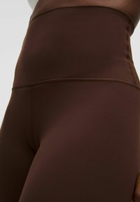 Leggings marron taille haute en tissu lisse et extensible avec des coutures plates et une coupe ajustée. Pas de logos ni de motifs visibles.