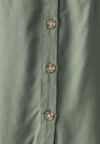 Gros plan sur une chemise en tissu vert avec trois boutons ronds translucides alignés verticalement, cousus avec un fil foncé.
