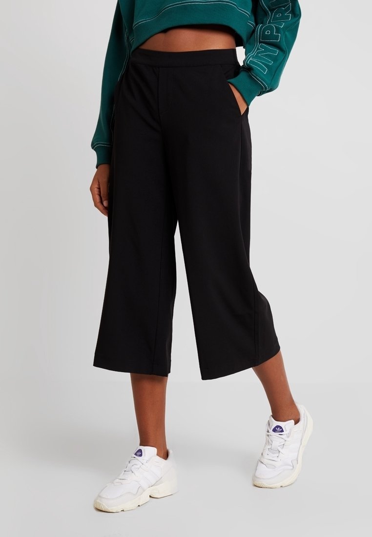 Pantalon noir à jambes larges avec une texture lisse, doté de poches avant et d'une taille haute, associé à des baskets blanches.