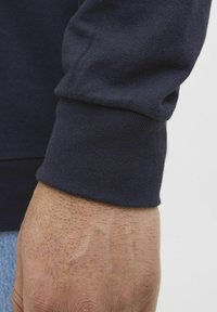Ärmelsaum eines marineblauen Sweatshirts mit gerippter Textur, eng anliegendem Schnitt und sichtbaren Nahtdetails. Der Hautton der Hand am Rand des Ärmels.