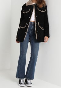 Svart mockajacka med fuskpälskrage och texturerad trim, med tryckknappar och två framfickor. Bärs ihop med utsvängda jeans och canvas-sneakers.