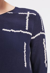 Laivansininen vaate, jossa on pehmeä rakenne ja valkoisia abstrakteja kuvioita. Suunnittelussa on risteäviä viivoja ja epäsäännöllisiä muotoja.