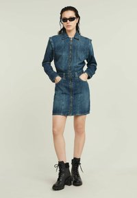 Denim jacket dress in dark blue, featuring a front zipper, collar, belted waist, and two side pockets. Complemented by black lace-up heeled boots.