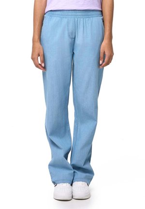 Un mannequin se tient devant un fond blanc, vêtu d’un pantalon bleu clair à jambes larges et à taille élastiquée, ainsi que de baskets blanches.