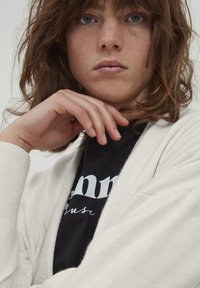 Strick-Cardigan in Creme mit offenem Vorderteil, gerippten Bündchen und weicher Textur. Schwarzes T-Shirt darunter mit weißem Text, Hände stützend auf dem Kinn.