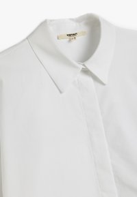 Chemise à col blanc en tissu lisse, dotée d'une patte de boutonnage et d'une étiquette indiquant "Koton Business", avec un design épuré et minimaliste.