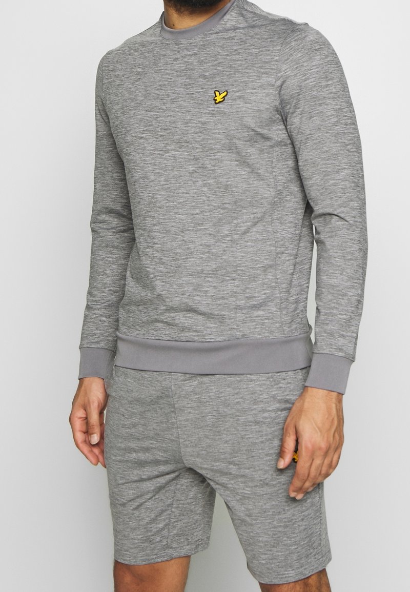Sweat-shirt gris à col rond avec tissu texturé, arborant un petit logo jaune sur la poitrine gauche. Associé à un short gris assorti.
