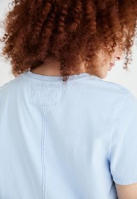 Lysblå t-shirt med rund hals, korte ærmer og en iøjnefaldende rygsøm detalje med et broderet mærke og synlige stikninger.
