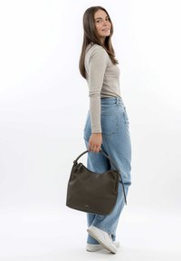 Sac à main en cuir vert foncé avec une finition douce et texturée, doté d'une anse courbée, tenu par une personne portant un jean clair et un haut beige.