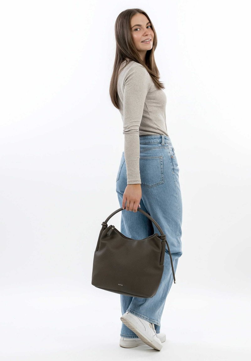 Sac à main en cuir vert foncé avec une finition douce et texturée, doté d'une anse courbée, tenu par une personne portant un jean clair et un haut beige.