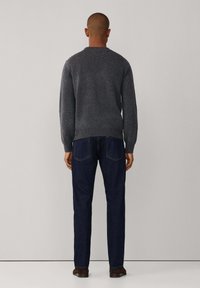 Grijze gebreide trui met ribbels aan de onderkant, gecombineerd met een donkere spijkerbroek. De achteraanzicht toont een gladde textuur en een eenvoudig ontwerp. Bruine schoenen zichtbaar.