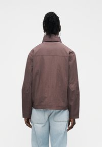 Giacca marrone leggera con collo alto e vestibilità ampia. Presenta spalle scese e una texture liscia. Indossata con jeans azzurri.