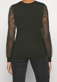 Donkergroen sweater met lange, transparante kantmouwen. Getailleerd ontwerp, geribbelde manchetten en zoom. Textuurpatroon van kant op de mouwen voegt detail toe.