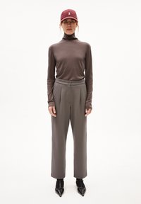 Langsleeve, taupe coltrui gecombineerd met lichtgrijze, high-waisted broek. Zwarte puntige schoenen en een bordeauxrode pet met logo.