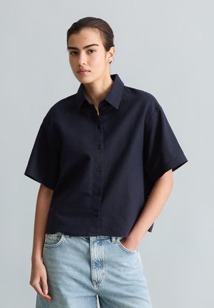 Młoda osoba ubrana w luźną, czarną koszulę z krótkim rękawem zapinaną na guziki oraz jasno-niebieskie jeansy, stojąca z jedną ręką w kieszeni.