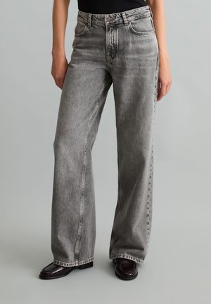 Personne portant un jean gris taille haute à jambes larges avec des chaussures noires et un haut sombre, debout sur un fond uni gris clair.