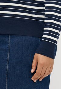 Ναυτικό και λευκό ριγέ πλεκτό πουλόβερ με ριχτές μανσέτες, συνδυασμένο με τζιν φούσ�τα. Το χέρι ακουμπά ελαφρά στην μέση της φούστας.