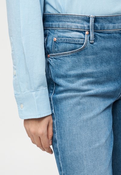 Osoba v svetlomodrej košeli a modrých rifliach, ruka spočíva blízko stehna, ukazujúc detaily vrecka na rifliach.