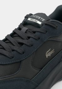 Le sneakers Lacoste presentano una tomaia di camoscio e materiali sintetici di un blu navy scuro, con lacci neri piatti e un'etichetta con il logo argentato. Accenti testurizzati presenti.