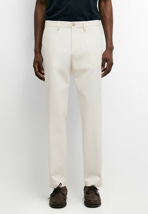 Homme portant un pantalon droit écru avec des passants de ceinture et des chaussures marron à lacets, debout sur fond blanc.