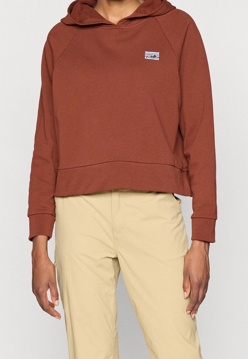 Personne portant un sweat à capuche couleur rouille avec un petit logo sur la poitrine et un pantalon beige, debout devant un fond uni.